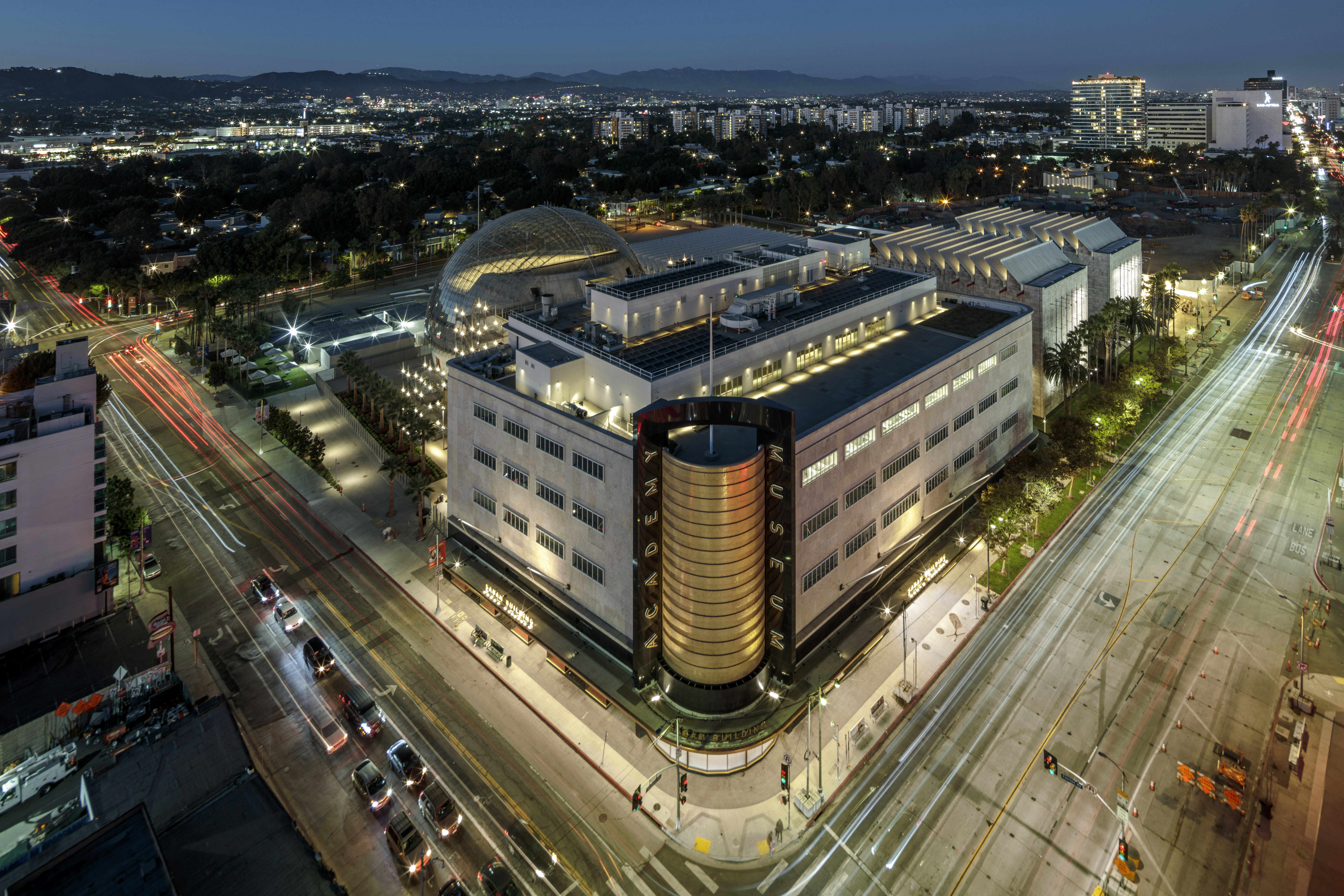 An aerial view photograph of a brightly lit building that takes up an entire city block. A gold column goes up the front in the center of the building.