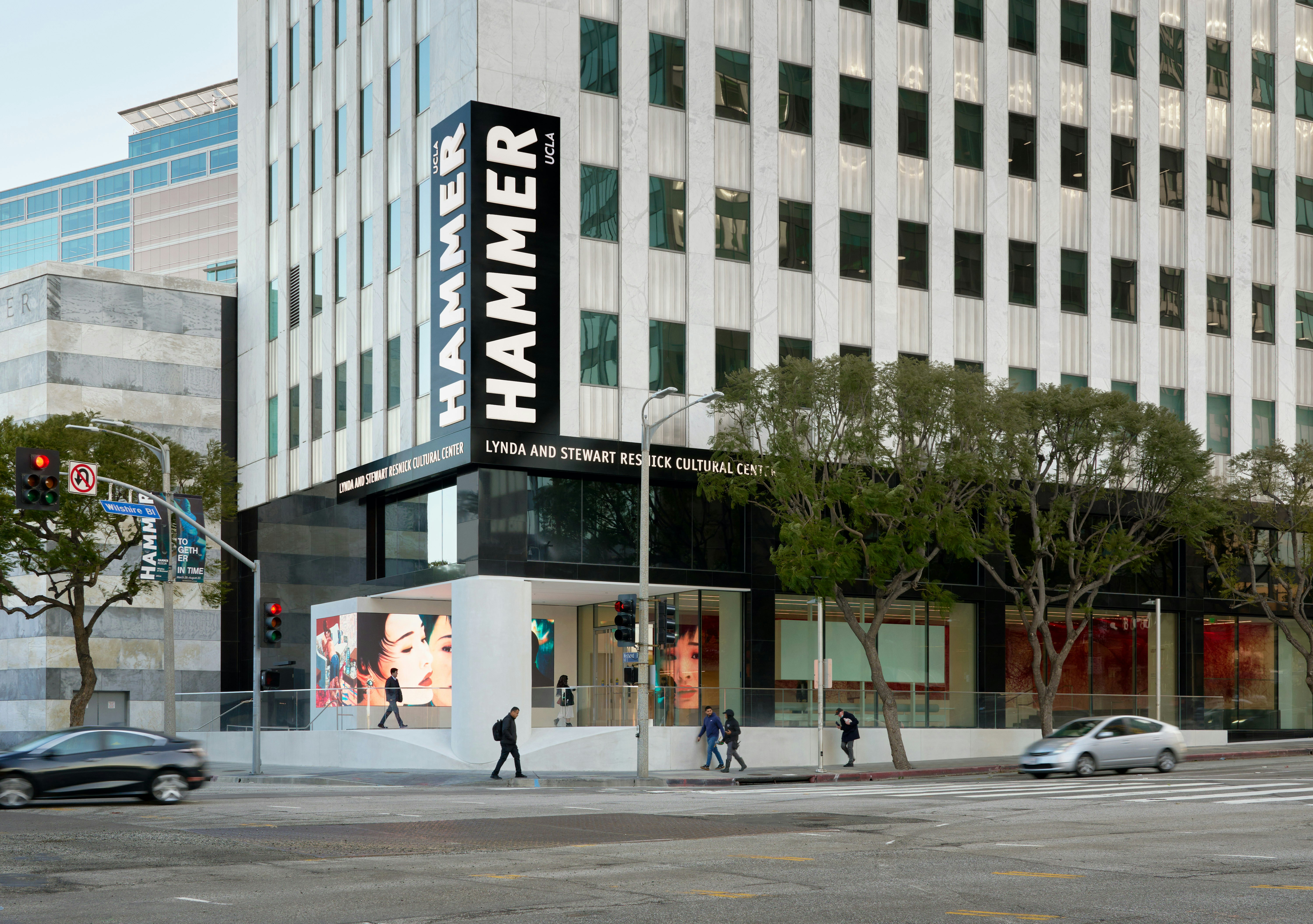 Exterior view of building with signage that reads: “Hammer UCLA” “Lynda and Stewart Resnick Cultural Center.” Moving cars trail across the main street, with museum banners on street poles.